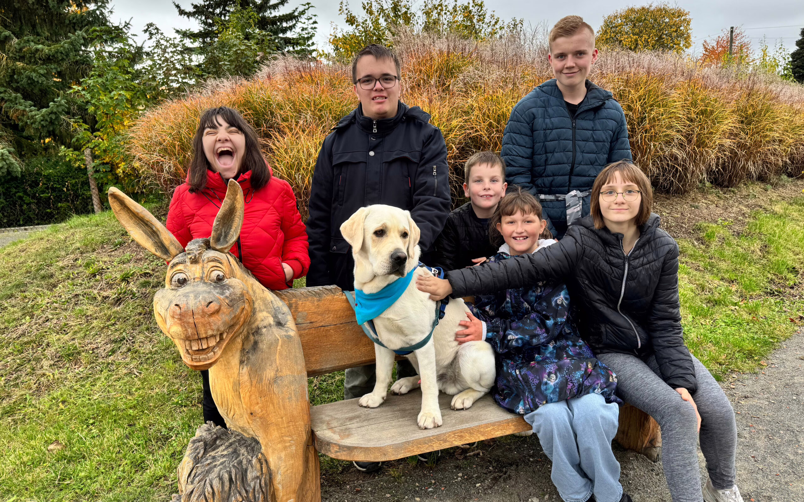 Sieben Kinder sitzen und stehen um eine geschnitzte Holzbank mit Eselsfigur; in der Mitte sitzt ein heller Labrador mit blauem Halstuch. Die Kinder lachen und schauen zur Kamera, im Hintergrund herbstliche Gräser und Bäume.